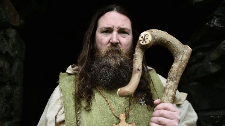 a man with long dark hair and beard is wearing a white and green robe and holding a shepherd's crook. he is a actor portraying saint patrick